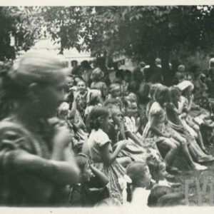 teren porośnięty trawą i drzewami, na którym siedzą i stoją ludzie w letnich ubraniach