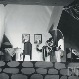 scena wewnątrz domu lisa, przy kuchence stoi lalka lisa ubrana w długą spódnicę i żakiet, miesza łyżką w garnku, po lewej stronie stoi stół z dwoma krzesłami, po prawej stronie łóżko, na ścianie wiszą dwa portrety lisów