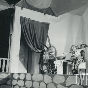 scena wewnątrz domku lisa, dwa zajączki siedzą przy stole, jedzą drewnianymi łyżkami z jednej miski, po lewej stronie drzwi wejściowe z kotarą