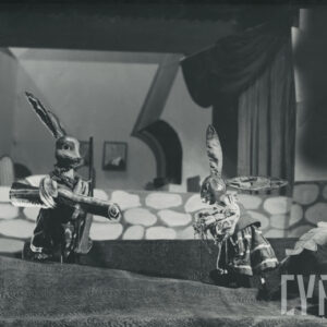 scena przed domkiem, dwie lalki zajęcy stoją naprzeciw siebie, jedna trzyma w ręku rulon materiału, na ramieniu ma strzelbę, druga trzyma w rękach pęczek włoszczyzny  