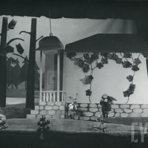 scena przed domkiem, naprzeciw siebie stoją dwie lalki zajęcy, za nimi okazały domek ze spadzistym dachem, wieżyczkami, porośnięty bluszczem