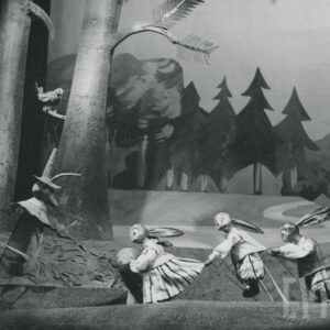 scena w lesie, po lewej stronie kukiełka świerszcza w kapeluszu ze skrzypcami w rękach, nad nim na drzewie siedzi ptak, obok jedna za drugą stoją kukiełki zajęcy, z tyłu ścieżka i las