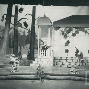 scena przed domkiem, na ganku stoi lalka zająca, celuje ze strzelby do dwóch lalek lisicy i wilka stojących po prawej stronie, po lewej stronie obok domu i za drzewami stoją lalki zajęcy