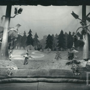 scena w lesie, pomiędzy drzewami stoi lalka przedstawiająca świerszcza w kapeluszu ze skrzypcami w ręku, po lewej stronie za drzewem stoi lalka przedstawiająca zająca, z tyłu ścieżka i las