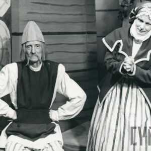 scena przed domem, mężczyzna z długimi wąsami ubrany w białą koszulę, kamizelkę oraz czapkę siedzi i podpiera się pod boki, obok stoi kobieta w spódnicy w pasy i marynarce z chustą na głowie, dłonie złożone jak do modlitwy