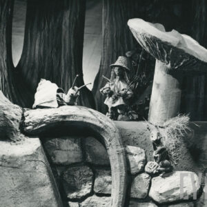 scena w lesie, wysokie drzewa, na wystającym korzeniu siedzi lalka przedstawiająca ślimaka, obok pod grzybem stoi lalka z długimi włosami w wiklinowym kapeluszu, trzyma łuk i strzały, poniżej na kamieniu wiewiórka 