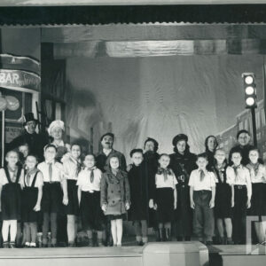 scena na ulicy przed barem mlecznym, w pierwszym rzędzie stoją elegancko ubrane dzieci w chustach na szyjach, w drugim rzędzie stoją dorośli aktorzy w kostiumach, po lewej stronie choinka