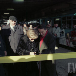 grupa osób stojąca na przystanku tramwajowym przy symbolicznej wstędze, którą kobieta przecina nożyczkami; w głębi stoi duża grupa osób przy tramwaju