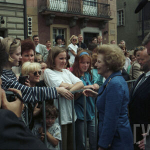 elegancka, starsza kobieta stojąca przy ogrodzeniu, za którym stoi duża grupa osób