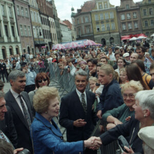 elegancka, starsza kobieta stojąca przy ogrodzeniu, za którym stoi duża grupa osób