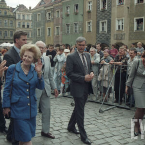 elegancka, starsza kobieta idąca po płycie rynku, otoczona przez elegancko ubranych mężczyzn, w tle za ogrodzeniem stoi duża grupa osób, widoczne kamienice