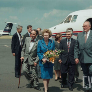 elegancka, starsza kobieta trzymają ca w dłoniach kwiaty, w towarzystwie starszych mężczyzn ubranych w garnitury; w tle m.in. inni ludzie i samolot
