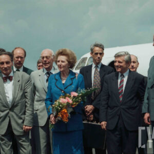 elegancka, starsza kobieta trzymająca w dłoniach kwiaty, w towarzystwie starszych mężczyzn ubranych w garnitury