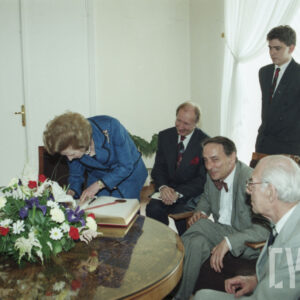 starsza kobieta wpisująca się do księgi wśród grupy starszych mężczyzn w eleganckich ubraniach siedzących przy okrągłym stole, na którym stoi wazon z kwiatami