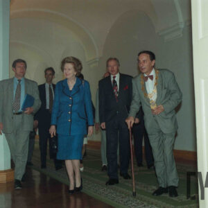 starsza, elegancko ubrana kobieta i grupa mężczyzn w garniturach w korytarzu budynku