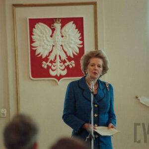 starsza, elegancka kobieta stojąca przed mikrofonem, za jej plecami na ścianie wisi godło narodowe