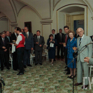 grupa odświętnie ubranych osób słuchająca przemówienia i trzymająca w rękach kieliszki z winem