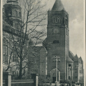 potężna wieża nakryta stożkowym hełmem, po lewej fragment dużego gmachu przysłonięty drzewem bez liści