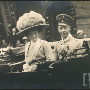 para siedząca w karocy, kobieta w ozdobionym tiulami kapeluszu z wielkim rondem, mężczyzna w mundurze galowym, w czapce i przy szabli