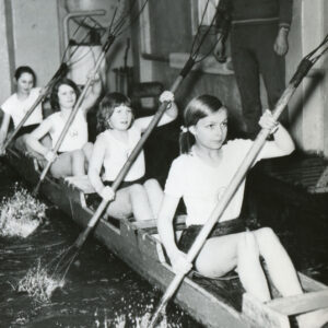 dzieci ubrane w stroje sportowe, trenujący w prowizorycznym kajaku i przy pomocy prowizorycznych wioseł