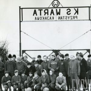 grupa osób w dresach sportowych, część z nich nosi czapki, pozująca do fotografii na tle bramy