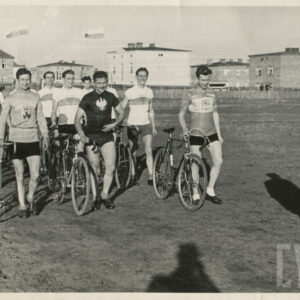 grupa mężczyzn w strojach sportowych, prowadzących rowery w czasie przemarszu przez trawiaste boisko