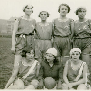 grupa kobiet w strojach sportowych, cztery z nich stoją, trzy siedzą, jedna z nich trzyma w rękach piłkę do gry; kobiety pozują do fotografii na trawiastym boisku