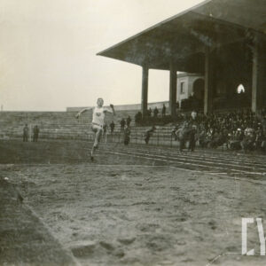 młody mężczyzna w stroju sportowym przygotowujący się do skoku na skoczni przy bieżni stadionu, w głębi trybuny stadionu, na których siedzą widzowie