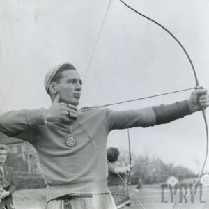 młody mężczyzna w stroju sportowym z herbem klubowym na piersi, mierzący do celu z łuku