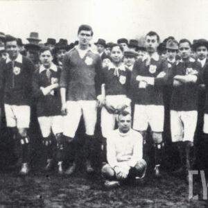 grupa młodych mężczyzn w strojach sportowych, jeden z nich siedzi, reszta stoi, pozująca do fotografii na tle zebranego tłumu 