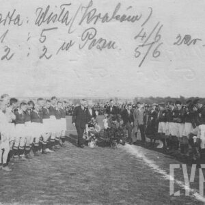 grupa mężczyzn w strojach sportowych, stojąca w dwóch szeregach naprzeciw siebie na boisku do piłki nożnej w towarzystwie  pięciu mężczyzn i jednej kobiety, noszących eleganckie ubrania; wokół trawiastego boiska stoją i siedzą widzowie