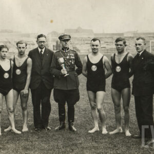 grupa mężczyzn stojących w szeregu na trawie; czterech z nich ubranych jest w stroje kąpielowe z herbem klubu na piersi, czterech kolejnych nosi garnitury; jeden mężczyzna w środku trzyma w ręku puchar i ubrany jest w mundur wojskowy 