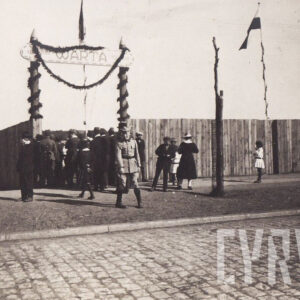 elegancko ubrani ludzie oraz oficer Wojska Polskiego przed wejściem na teren ogrodzony drewnianym płotem, nad wejściem przyozdobiony girlandą napis Warta