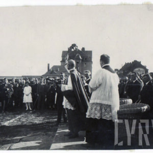 dwóch duchownych oraz duża grupa odświętnie ubranych ludzi stojąca na boisku sportowym