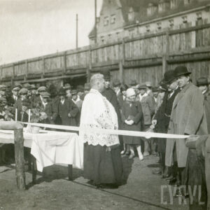 duchowny stojący wśród elegancko, odświętnie ubranych ludzi stojących przy wstędze na terenie boiska sportowego; w tle drewniany płot i budynek z cegieł