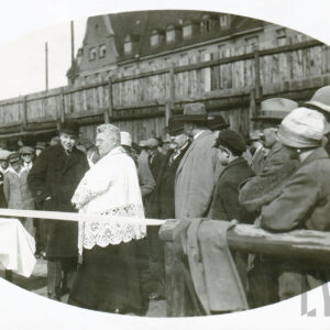 duchowny stojący wśród elegancko, odświętnie ubranych ludzi stojących przy wstędze na terenie boiska sportowego; w tle drewniany płot i budynek z cegieł