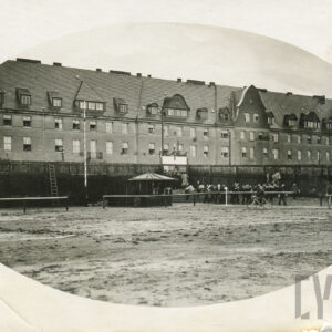 boisko sportowe otoczone drewnianym parkanem, w głębi ceglany budynek mieszkalny