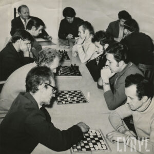 grupa elegancko ubranych mężczyzn siedząca przy stole i pochylona nad szachownicą