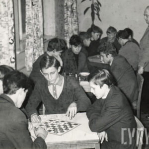 grupa elegancko ubranych mężczyzn siedząca przy stole i pochylona nad szachownicą