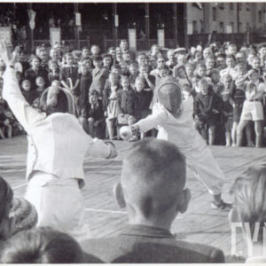 dwóch mężczyzn w strojach szermierczych i floretami w dłoniach walczący na planszy ustawionej na murawie stadionu, otoczonej grupą widzów