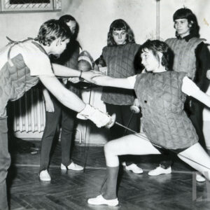 młode kobiety w strojach ochronnych, wśród nich stoi mężczyzna poprawiający pozycję szermierczą jednej z zawodniczek 