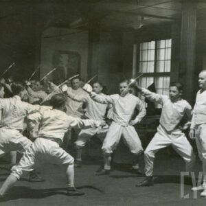 grupa mężczyzn w strojach szermierczych stojących w dwóch rzędach naprzeciw siebie i trenujących uderzenia floretem