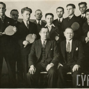 grupa mężczyzn w garniturach pozująca do fotografii, dwóch starszych siedzi na ławce, pozostali stoją za nimi, a w rękach trzymają rakietki do tenisa stołowego