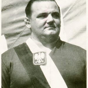 potężny mężczyzna ubrany w dres sportowy, przepasany biało-czerwoną wstęgą z godłem państwowym, pozujący do fotografii