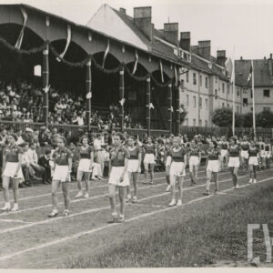 grupa kobiet w strojach sportowych maszerujących na bieżni sportowej przy trybunie stadionu, na której siedzą widzowie; w głębi budynki mieszkalne z cegły