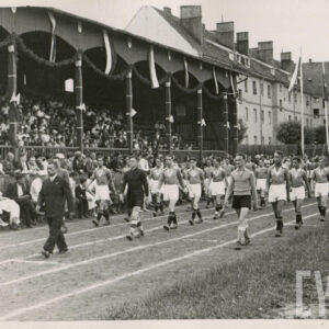 grupa mężczyzn w strojach sportowych maszerujących na bieżni sportowej przy trybunie stadionu, na której siedzą widzowie; w głębi budynki mieszkalne z cegły