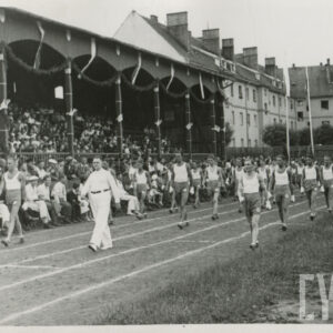 grupa mężczyzn w strojach sportowych maszerujących na bieżni sportowej przy trybunie stadionu, na której siedzą widzowie; w głębi budynki mieszkalne z cegły