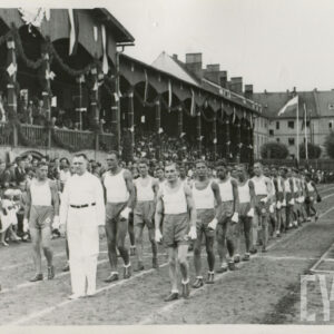 grupa mężczyzn w strojach sportowych maszerujących na bieżni sportowej przy trybunie stadionu, na której siedzą widzowie; w głębi budynki mieszkalne z cegły