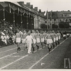 grupa mężczyzn w strojach sportowych maszerujących na bieżni sportowej przy trybunie stadionu, na której siedzą widzowie; w głębi budynki mieszkalne z cegły