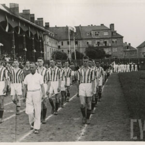 grupa mężczyzn w strojach sportowych maszerujących na bieżni sportowej przy trybunie stadionu, na której siedzą widzowie; w głębi budynki mieszkalne z cegły